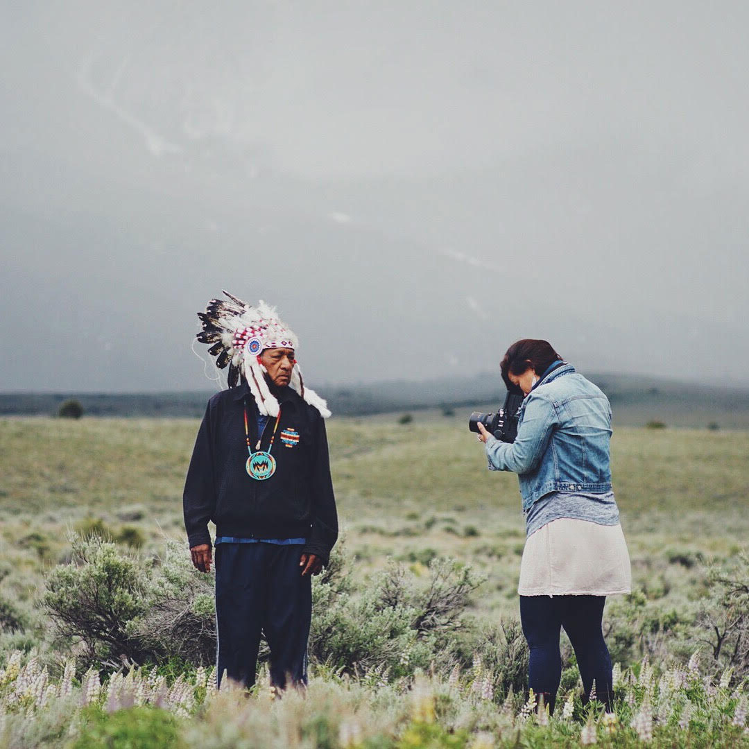 Matika Wilbur in a field at the foot of a mountain range taking a photograph of a native american man in full headdress