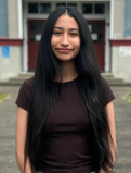Ariana smiles for the camera in a headshot. She's wearing a plain dark brown T-shirt, and stands infront her highschool entrance. 