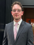 Blayne stands in a suit with a pink checkered tie, he looks to be in a theater. 