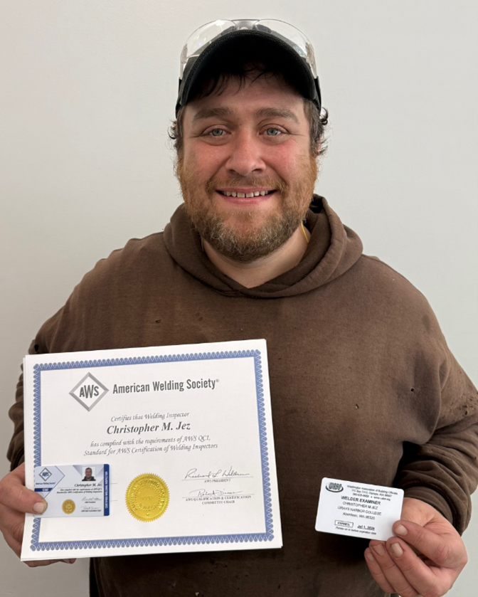 Wearing a brown pull over hoodie and baseball cap, Chris Jez proudly present his certificates from the american welding society to the camera, a smile on his face.  In his right hand is his new Welding Inspector certification, and an illegible card, in his left is his welding examiner ID card. 