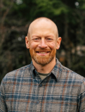 Kevin grins for the camera in a professional Headshot. He wears a grey, orange, and blue plaid flannel, with a contrasting collar underneath.