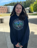 Madeline stands for a photo, hands clapsed infront of her, on the GHC campus. She is wearing a black hoodie with a circular logo on it. 