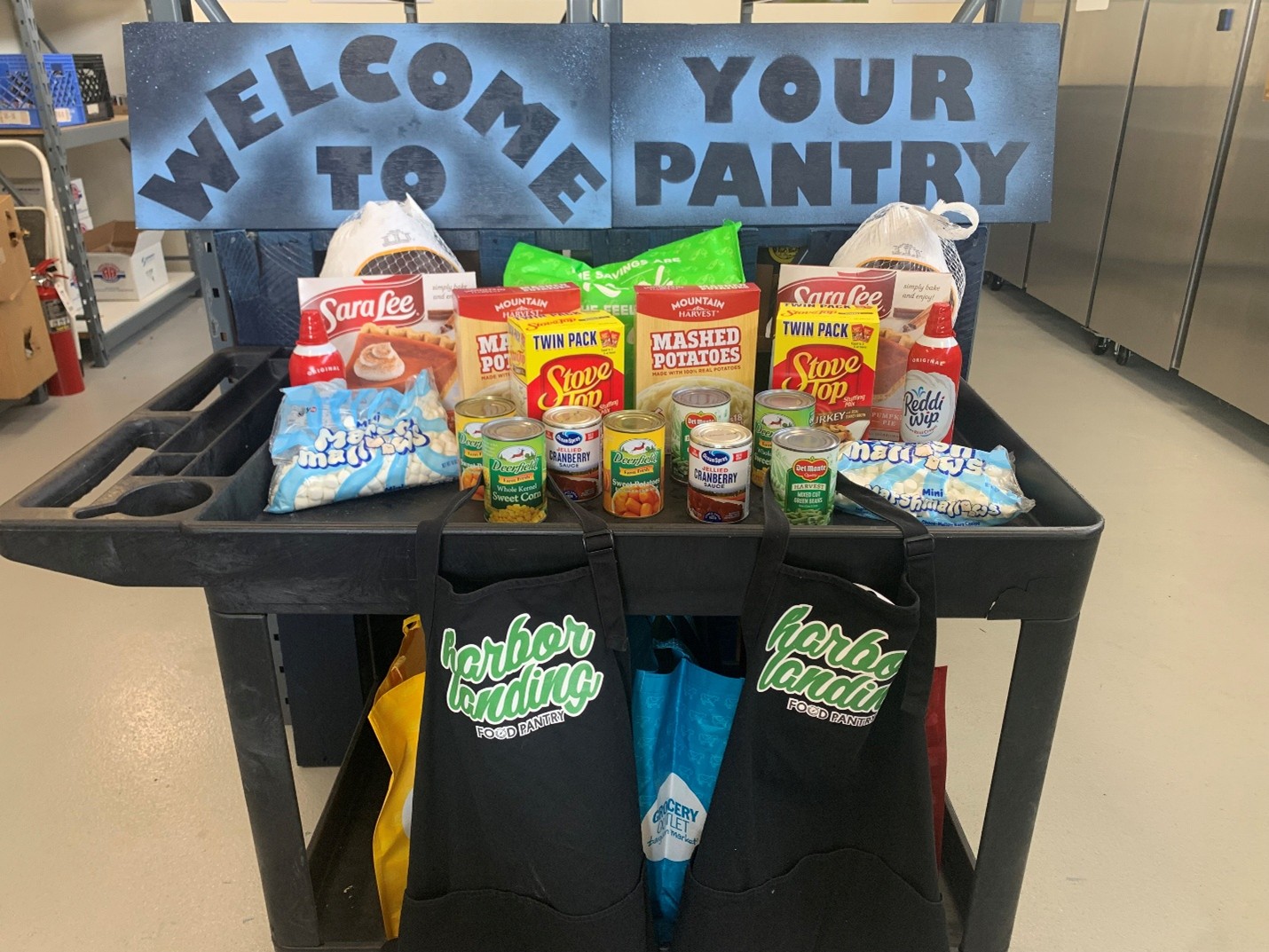 A black cart with varied Thanksgiving typical food on it. Visible are marshmellows, pumpkin pie, instant mash potatoes, stovetop turkey stuffing mix, packaged turkeys, canned sweet potatoes, corn, and cranberry sauce. Above the food are spraypainted signs from the food pantry saying "Welcome to your pantry" And hanging off the front are  black aprons branded with the Harbor Landing Food Pantry logo, covering branded reuseable shopping bags. One from grocery outlets peaks out the gaps.. 