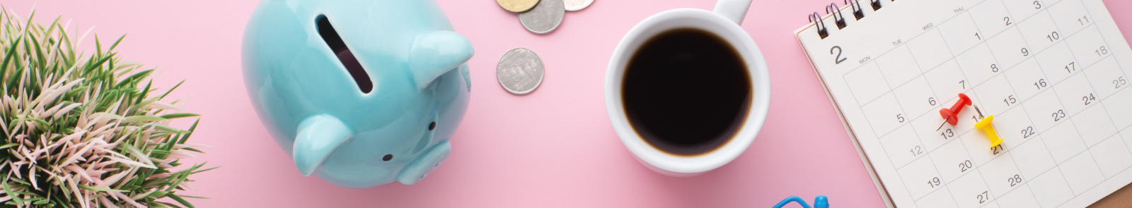 The camera looks from above at a pink desk space. From left to right is a varigated potted plant, a blue piggybank, loose change, a mug of coffee, and a calender with thumbtacks resting on the top as if about to be put up on the wall. 