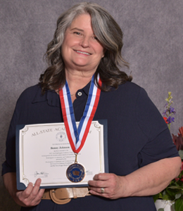 Bonnie smiles for the camera holding a paper reward. She is wearing a black polo shirt which makes the red white and blue lanyard of her medal pop. 