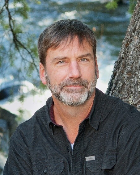 Derek Sheffield in a headshot. He wears a charcoal button up and a freshly trimmed salt and pepper beard and stache. The hair on the top of his head is whispy, with considerably less greying. In the background is an indistinct body of water, and a tree trunk as if he is sitting just infront of it.