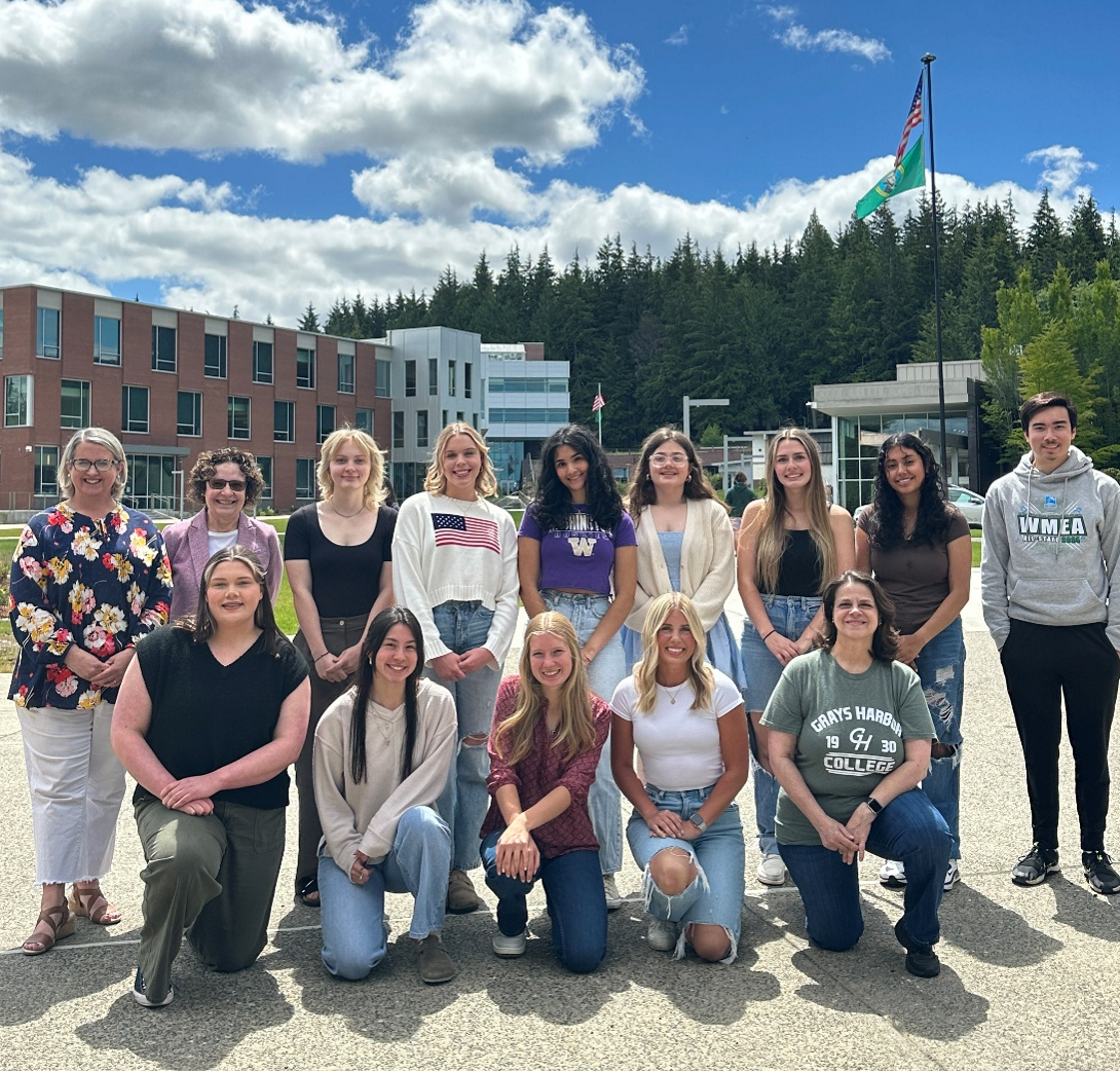 12 students and some staff smile for the camera in two rows, the bottom kneeling. The Library and Tulalw building are in the background and its a bright day with scattered clouds. 