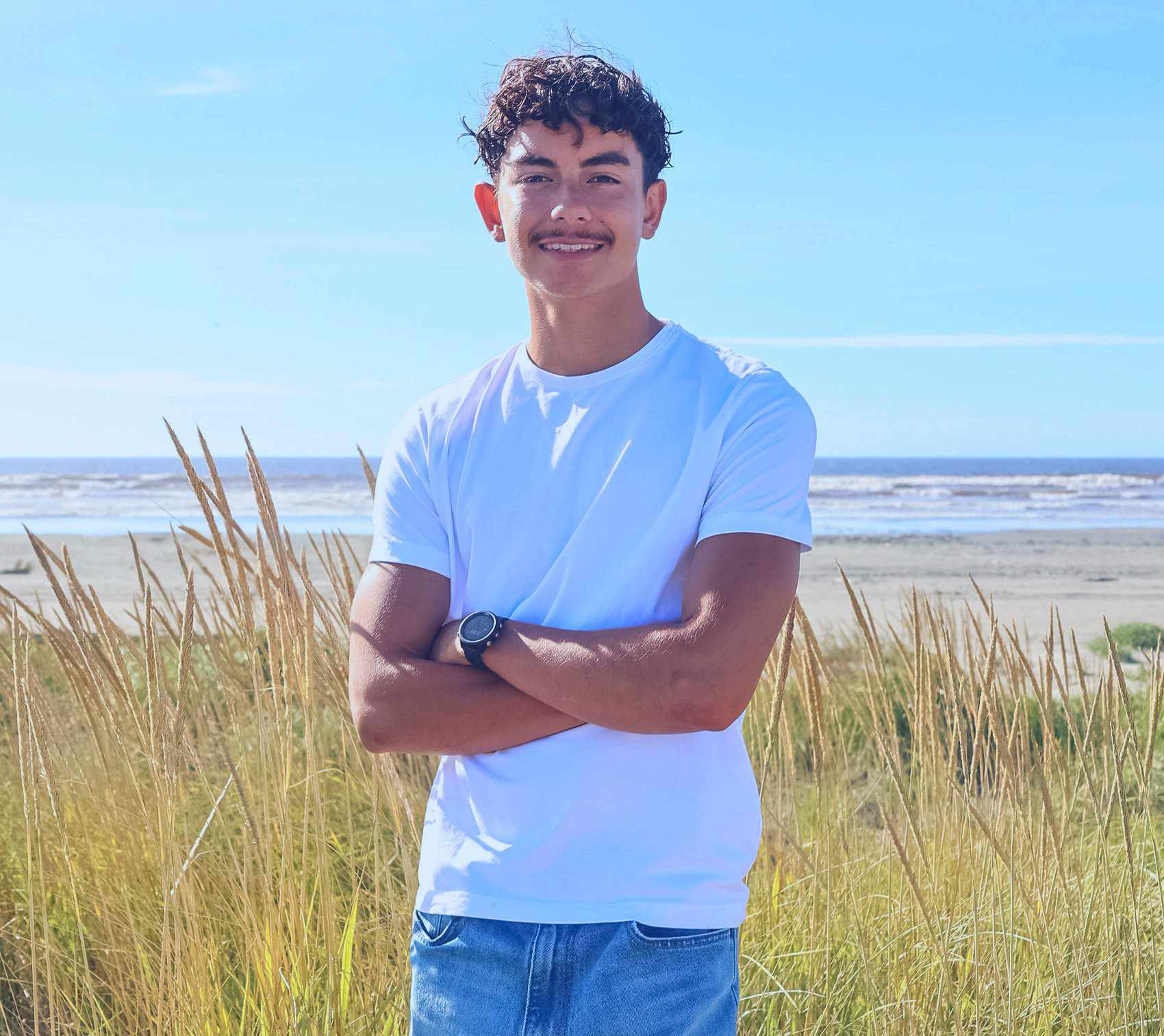 Noah Aquino smiles for the camera infront of a grassy beach shore. 