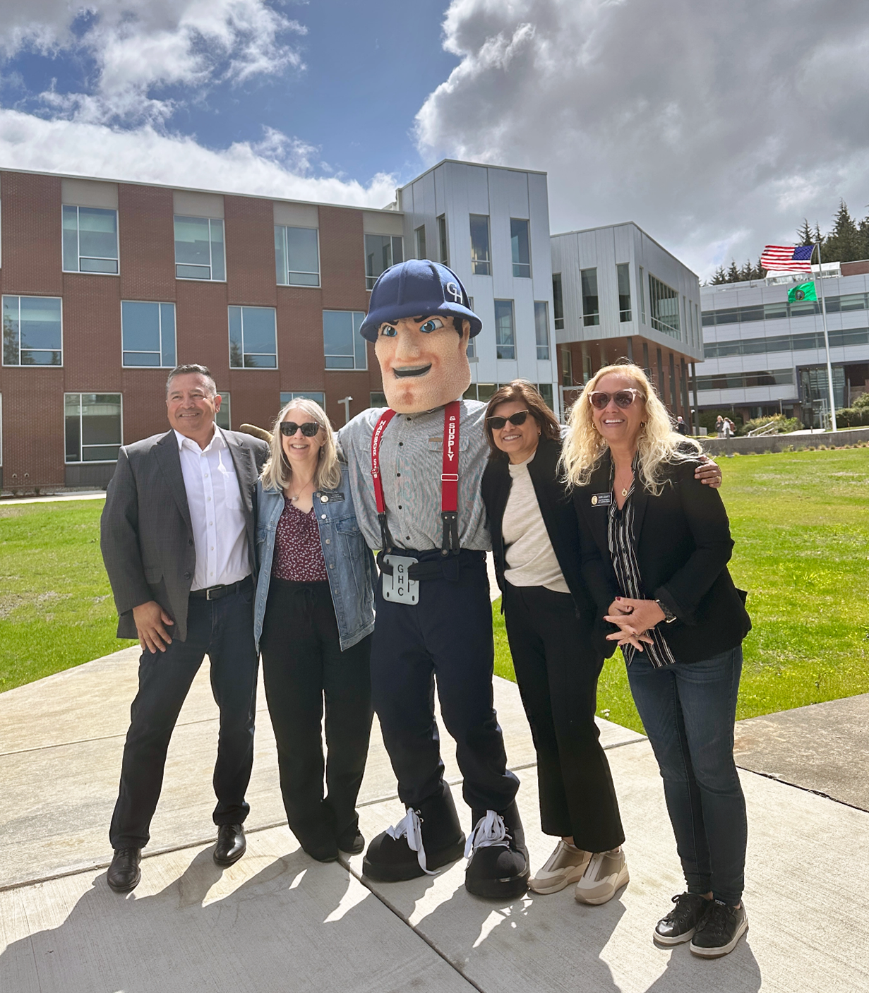Charlie Choker wraps his arms around the shoulders of four people dressed in business wear. They all smile towards the camera and three of them are wearing sunglasses to account for the bright day visible behind them.