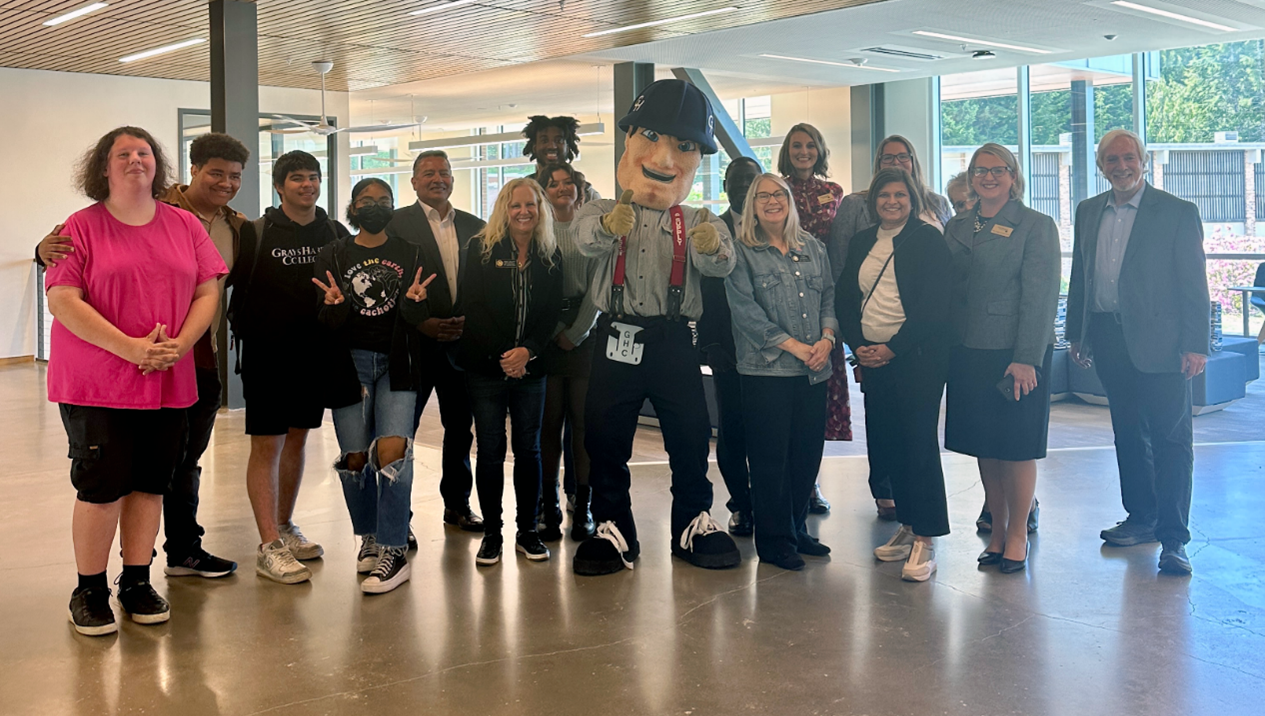 A group of 17 stand and smile for the camera in a brightly lit lounge space. Charlie Choker, GHC's mascot, stands front and center, giving two enthusiastic thumbs up to the camera.