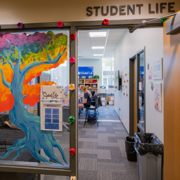 A door to an office with the words Student Life on top of it. A window with a painted blue, orange, green, and purple tree