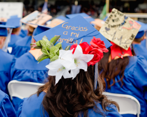 A student sitting at graduation wearing a graduation cap that has the words Gracias a mi Familia and some flowers on it