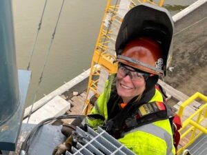 Tara crouches next to water on a job site, a piece of metal grating in hand. She's wearing a high vis vest and a welding mask, flipped up so the camera can see her grinning expression.
