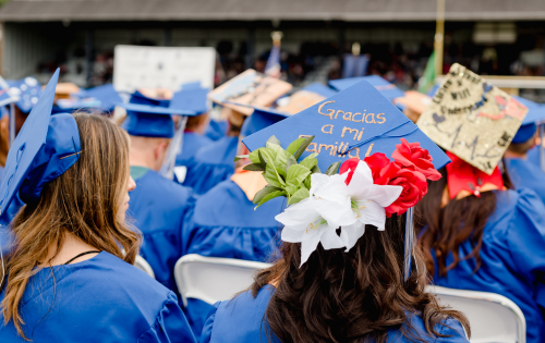 Students at commencement wearing graduation caps and gowns. The cap in focus says, “thank you to my family” in Spanish