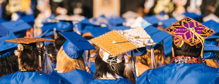Over the top of a photo of the back of graduates’ heads featuring their caps is a blue arrow with the words Tell Us How We Did!