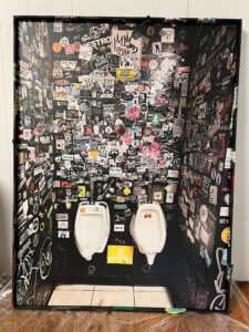 Two urinals of different make in a room side by side. The walls are poorly painted a harsh black to contrast with the white tile and porcelain. Nearly all of the walls are covered in various stickers and advertisements. Some graffiti tags lower on the makeshift canvas.