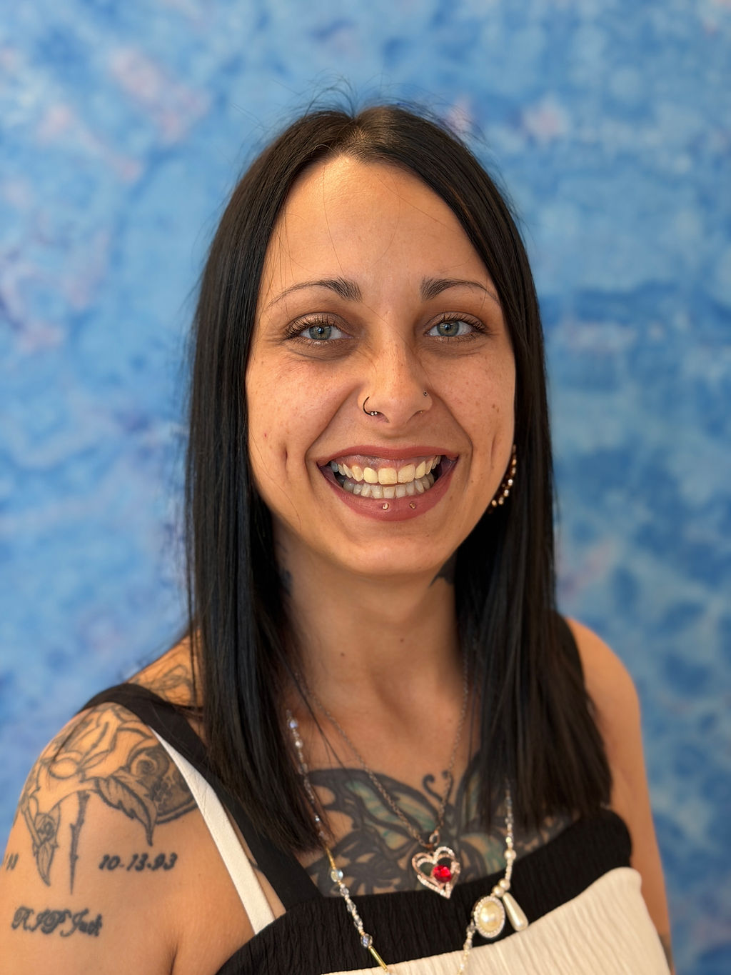 Scyland smiles for the camera in a professional photo. Scyland has two bottom lip piercings, a nostril ring, and large studded hoop earrings that peak out from dark hair. 
